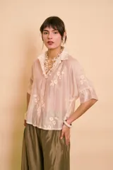 Camisa de manga corta color beige, translúcida, con cuello camisero, cierre frontal con botones y bordados florales tono sobre tono.