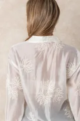 Camisa de manga larga en color blanco, confeccionada en tejido traslúcido con delicados bordados florales en hilo de seda al tono. Presenta cuello clásico, cierre frontal con botones y puños abotonados.
