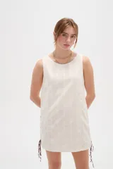 Vestido corto de algodón color blanco, de corte holgado, sin mangas y con cuello a la base. Presenta detalles de cordones cruzados y ojalillos en los laterales.