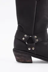 Bota de caña alta estilo western confeccionada en cuero vacuno negro. Presenta un diseño robusto con taco cuadrado de madera, punta redondeada y una tira decorativa con herrajes metálicos y tachas en la zona del tobillo.