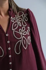 Blusa color bordó con bordados florales en tono crema en la parte superior frontal. Presenta cuello redondo con pequeña abertura, cierre frontal con botones y mangas largas acampanadas con puños ajustados.