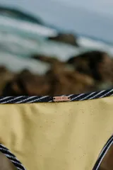 Bombacha de bikini negra con ribetes en color amarillo pastel. Presenta tiras laterales finas ajustables y una pequeña placa metálica decorativa con el nombre de la marca en la parte superior.