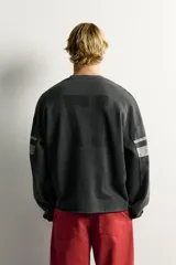 Sudadera cropped fit de color negro con efecto lavado vintage, cuello en V y manga larga. Presenta estampado con texto "LOS ANGELES" y el número "17" en parches acolchados en el frente, además de bandas grises en las mangas.