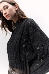 Sweater de lana merino tejido en punto grueso, color gris oscuro, con cuello alto. Presenta un diseño calado con tientos de cuero marrón insertados a lo largo de la prenda, simulando un lazo o detalle orgánico.