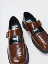 Zapato tipo guillermina de cuero color marrón con acabado brillante, punta cuadrada y detalle de costuras en relieve sobre el empeine. Cuenta con una correa ajustable con hebilla metálica y suela de goma negra con taco bajo.