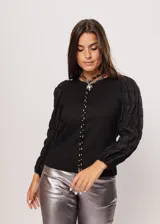 Blusa negra de tejido de punto acanalado, con cuello redondo y mangas largas abullonadas de tela semitransparente. Presenta una hilera vertical de pequeñas cuentas plateadas en el frente.