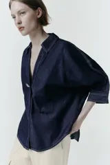 Camisa de denim azul oscuro con corte amplio, cuello solapa, escote en pico y mangas tres cuartos. Presenta un aplique metálico dorado en el centro y bajo asimétrico.