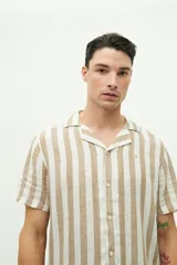 Camisa de manga corta con cuello cubano, confeccionada en una mezcla de lino y algodón con textura. Presenta un diseño a rayas verticales en tonos beige y celeste sobre fondo blanco, con cierre frontal de botones y logo bordado en el pecho.