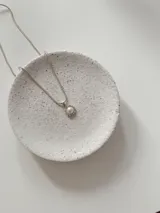 Collar de cadena fina tipo serpiente en tono plateado con un dije circular que presenta una perla central rodeada por pequeñas piedras brillantes.