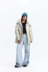 Campera puffer larga con capucha, color beige, de ajuste relajado. Presenta cierre frontal con botones a presión, bolsillos laterales con cierre y puños tejidos acanalados.