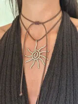 Collar con dije de sol estilizado en metal plateado, con centro en espiral y rayos rectos. El dije cuelga de un tiento sintético trenzado de color marrón oscuro, ajustable mediante un nudo corredizo.