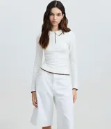 Blusa de punto color blanco marfil con cuello polo y cierre frontal de botones. Presenta ribetes en tono marrón oscuro en el cuello, los puños y el dobladillo inferior, con mangas largas y diseño ajustado.