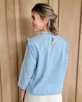 Blusa blanca de manga larga con cuello alto y volados en cuello y hombros.