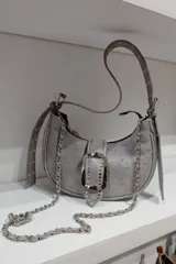 Cartera bandolera de cuero vacuno color gris con textura granulada. Tiene correa de hombro ajustable y extraíble, cierre superior con cremallera y herrajes metálicos plateados. El frente está adornado con una hebilla rectangular con incrustaciones de strass.