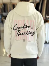 Canguro oversize de color beige claro con capucha, mangas largas y puños elastizados. Presenta un diseño gráfico en la espalda con el texto 'Creative Thinking' en tipografía cursiva negra sobre un fondo de letras grandes en tono rosado.