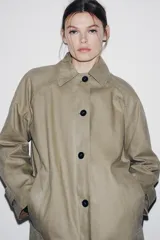 Trench coat de corte largo y diseño minimalista, confeccionado en tejido encerado color beige. Presenta cuello solapa, cierre frontal con botones oscuros y bolsillos laterales integrados.