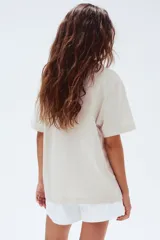 Remera de punto de algodón color beige claro, con cuello redondo y manga corta. Presenta un estampado gráfico en negro que incluye tres palmeras, un tigre caminando y el texto "COASTAL" y "OASIS" enmarcado.