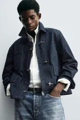 Chaqueta de denim de algodón en color azul oscuro, con cuello solapa y manga larga. Presenta cierre frontal con cremallera oculta bajo una solapa con alamares metálicos, bolsillo de vivo en el pecho y bolsillos de plastrón con solapa en la cadera.