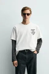 Camiseta blanca de corte relajado, con cuello redondo y manga corta. Presenta un pequeño estampado de texto en contraste en la parte frontal izquierda del pecho.
