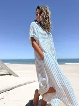 Vestido camisero midi de corte holgado, con estampado de rayas verticales celestes sobre fondo blanco. Presenta cuello camisero, cierre frontal con botones, mangas cortas con detalle de trabilla y bolsillos laterales.