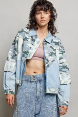 Chaqueta de denim de corte holgado y manga larga, con diseño en técnica de patchwork que combina sectores de mezclilla clásica, estampa de camuflaje, parches florales y detalles de encaje blanco. Cuenta con cierre frontal de cremallera metálica y cuello camisero.
