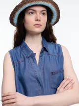 Camisa sin mangas en denim 100% lyocell, muy liviano con caída fluida. De corte recto y largo medio, presenta cartera escondida en el frente y un bolsillo plaqué en el pecho. En la espalda, un detalle distintivo: botones decorativos alineados en abertura vertical. Los pespuntes a contratono completan el look.