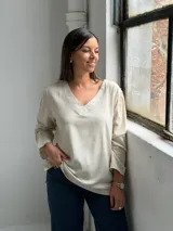 Blusa de lino color beige con escote en V, mangas largas con sutiles frunces en los hombros y costuras decorativas visibles en el cuello y el ruedo.