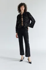 Pantalón de denim negro con diseño de tachas metálicas distribuidas en patrones curvos. Presenta un corte recto y tiro medio.