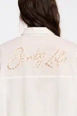 Camisa blanca de corte recto con cierre de botonadura frontal y cuello camisero. Manga larga con puños y botón, bolsillo de plastrón en el pecho y maxicanesú en espalda con logo bordado.