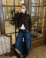 Campera de estilo militar en color marrón oscuro, confeccionada en gamuza. Presenta un diseño corto, cuello alto y cierre frontal cruzado con botones metálicos decorativos. Incluye detalles de charreteras en los hombros y botones en los puños.