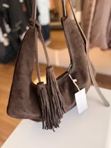 Cartera tipo hobo de textura gamuzada en color marrón oscuro, con diseño flexible y asa larga regulable. Presenta un detalle de flecos largos desmontables en uno de los laterales y cierre superior.