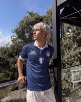 Camiseta de fútbol de Escocia de manga corta, color azul marino con diseño a cuadros sutiles, cuello en V y cuello camisero blanco con líneas azules. Presenta detalles estampados en el frente, incluyendo el escudo de la Asociación de Fútbol de Escocia, un león rampante y el texto 'Scottish Football Association'.