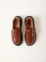 Mocasines de cuero color marrón con plataforma dentada negra. Presentan una tira frontal con hebilla metálica dorada y costuras decorativas en contraste.