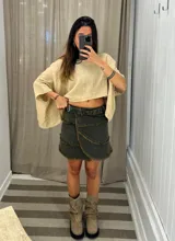 Minifalda cruzada tipo portafolio de denim color verde oscuro/gris, con bordes deshilachados en capas superpuestas y ajuste lateral con hebilla y ojalillos metálicos.