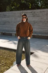 Campera de gamuza color marrón con acabado cepillado suave, cuello clásico, hombros caídos y mangas largas. Cuenta con cierre frontal oculto bajo solapa con botones a presión, bolsillos laterales con cierre y diseño abullonado con elástico interior en el bajo.