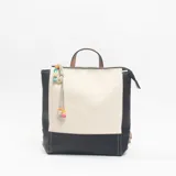 Mochila de diseño bicolor en negro y beige, con cierre superior, asa de mano y detalle de colgante con cuentas de colores en el lateral.