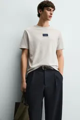 Camiseta de corte relajado, color gris jaspeado, con cuello redondo y manga corta. Presenta un parche rectangular azul marino en el centro del pecho con texto estampado en blanco y una frase adicional debajo.
