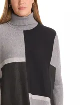 Polera tipo poncho de corte oversize, con cuello alto tejido y mangas largas con puños elásticos. Presenta un diseño geométrico de bloques de color en tonos beige, gris y tostado.