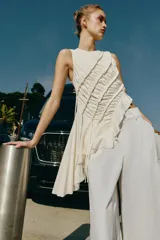 Top de algodón color crudo, con diseño sin mangas, cuello redondo y bajo asimétrico acabado en volante. Presenta frunces diagonales con detalles de cordones y cierre trasero con abertura y botón.