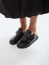 Mules de cuero negro con diseño de punta redondeada, suela plana y tira ajustable con hebilla metálica sobre el empeine, destacada por costuras a contratono.