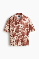 Camisa de manga corta con cuello abierto, tapeta de botones y bajo recto. Presenta un diseño estampado con motivos florales en tonos tierra y blanco.