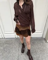 Minifalda de gamuza sintética en tonos marrones, con diseño en capas asimétricas, detalles de tachas metálicas doradas en los bordes y cierre cruzado con broches a presión.