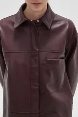 Sobrecamisa de cuero sintético en tono bordó oscuro, con cuello clásico, cierre frontal oculto y un bolsillo aplicado en el pecho. Presenta un corte holgado con mangas largas y puños rectos.