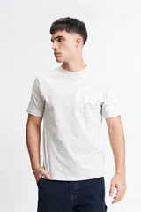 Remera de manga corta color beige claro, con escote redondo y fit regular. Presenta un estampado frontal con letras góticas en blanco.