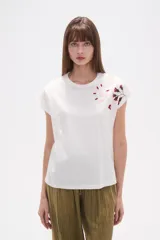 Remera blanca de algodón con mangas cortas y cuello redondo. Presenta un bordado floral en tonos rosados y rojizos en la zona del hombro derecho.