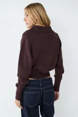 Sweater de punto color marrón oscuro, con cuello polo y cierre frontal parcial. Presenta mangas largas con puños anchos acanalados y un acabado acanalado en el bajo.