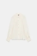 Blusa fluida de color blanco roto o crema, con cuello alto fruncido y manga larga con puños elásticos tipo nido de abeja. Presenta un frente abotonado con botones pequeños y brillantes.