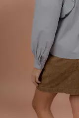 Blusa de jeans fino color celeste con detalles de terciopelo negro en contraste. Presenta cuello alto con volados, mangas largas abullonadas con puños ajustados y volados decorativos en los hombros y el frente.