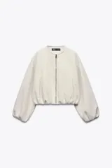 Chaqueta bomber de corte holgado y diseño minimalista, confeccionada en tejido suave color beige. Presenta cuello redondo, cierre frontal con cremallera metálica, mangas largas abullonadas con puños elásticos y bolsillos delanteros de vivo.