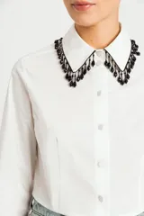 Camisa blanca de corte crop, con cuello clásico adornado con pedrería negra, cierre frontal de botones y mangas largas con puños abotonados.